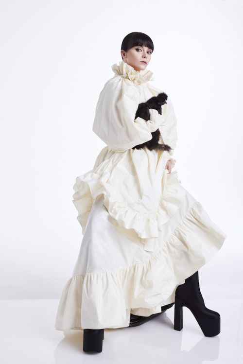 Woman in ruffled dress poses with a black object in a white photo studio, wearing platform boots and makeup for a fashion shoot.