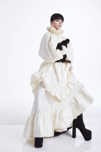Woman in ruffled dress poses with a black object in a white photo studio, wearing platform boots and makeup for a fashion shoot.