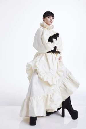 Woman in ruffled dress poses with a black object in a white photo studio, wearing platform boots and makeup for a fashion shoot.
