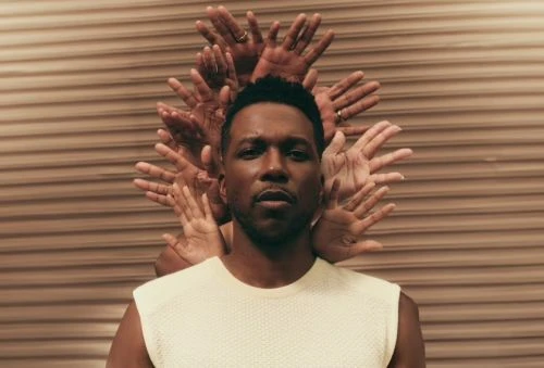 Portrait of a Black man wearing a cream sleeveless top, framed by multiple arms and hands against a tan horizontal-lined backdrop.