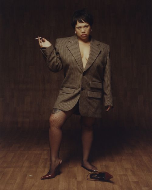 Woman in oversized blazer and fishnet stockings poses in an indoor setting. She's holding a cigarette and wearing pointed-toe heels.