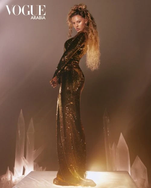 Woman in sparkling gold gown poses on a white platform with crystal formations against a light brown background, Vogue Arabia logo.