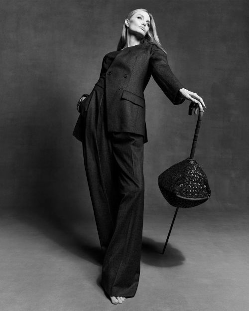 Blonde woman in dark double-breasted suit with wide-leg trousers poses in a photo studio with an ornamental bucket. Fashion editorial.