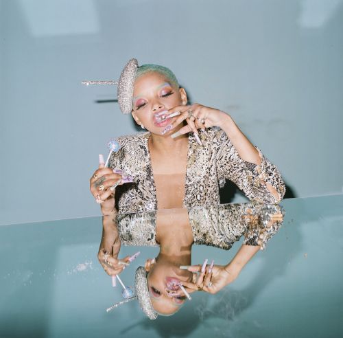 Unique close-up of Willow Smith with striking manicure and embellished headwear, captured in a light blue studio environment with reflection.