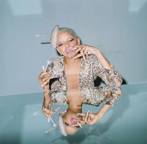 Unique close-up of Willow Smith with striking manicure and embellished headwear, captured in a light blue studio environment with reflection.