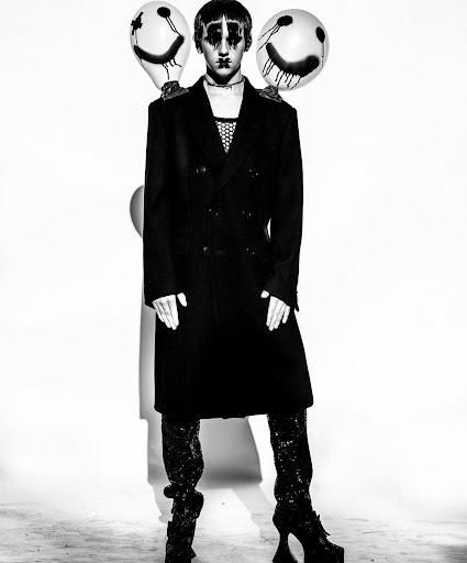 Person in dark avant-garde fashion wearing clown makeup and holding balloons with smiley faces against a white background.