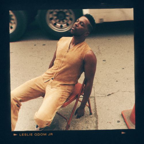 An adult male in a tan sleeveless outfit is relaxing on a red metal chair on a city sidewalk, with a utility truck in the background.