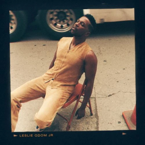 An adult male in a tan sleeveless outfit is relaxing on a red metal chair on a city sidewalk, with a utility truck in the background.