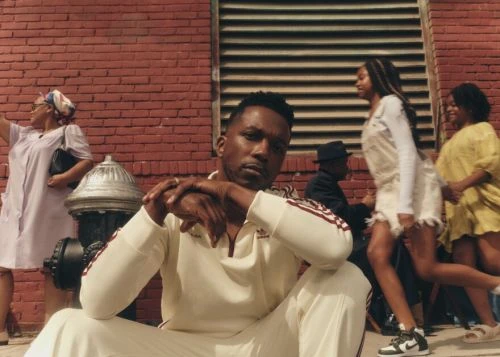 Stylish man in a cream Adidas tracksuit sitting with people in the background, urban scene, brick wall, and fire hydrant.