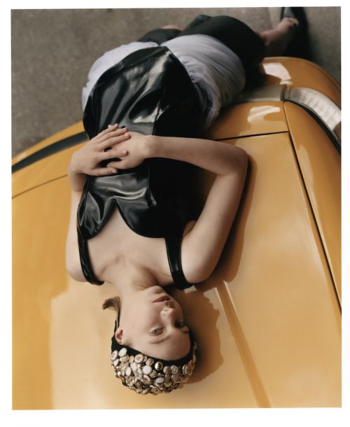 Eye-level portrait of a woman in a black dress lying on a yellow car, looking at the camera with a neutral expression and hands clasped.