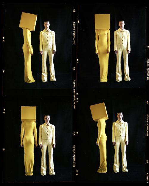 Two women in yellow outfits, one with a square box over her head, pose against a black background in a studio setting, editorial fashion.