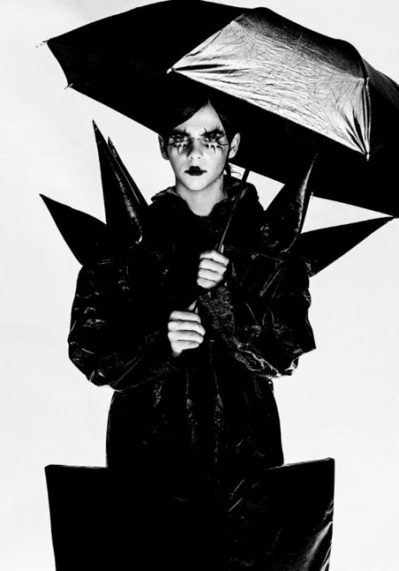 Person in dark makeup and avant-garde black fashion holding an umbrella against a white background in a studio setting.