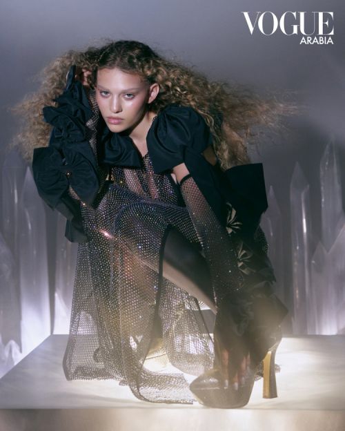 Fashion editorial shot: A woman with curly hair, wearing a jeweled dress and platform heels, poses against a gray background for Vogue Arabia.