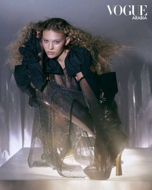 Fashion editorial shot: A woman with curly hair, wearing a jeweled dress and platform heels, poses against a gray background for Vogue Arabia.