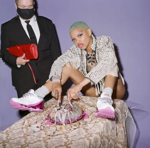 Stylish woman with buzzcut in embellished jacket poses near bouquet and man with red clutch against purple backdrop in a studio setting.