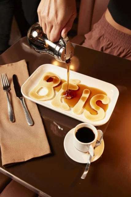 Close-up on pancakes spelling "Bois" on a white plate, syrup being poured on them. A cup of coffee and a set of silverware is also visible.