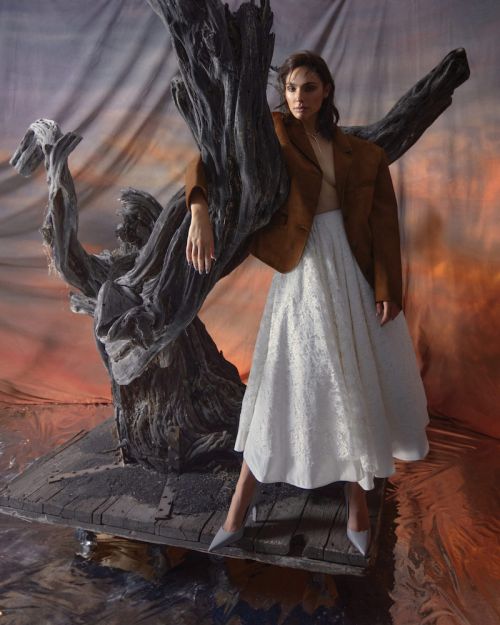 Brunette woman in white skirt and brown blazer posing with driftwood sculpture in front of a painted sunset backdrop in a studio.