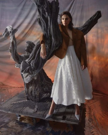 Brunette woman in white skirt and brown blazer posing with driftwood sculpture in front of a painted sunset backdrop in a studio.