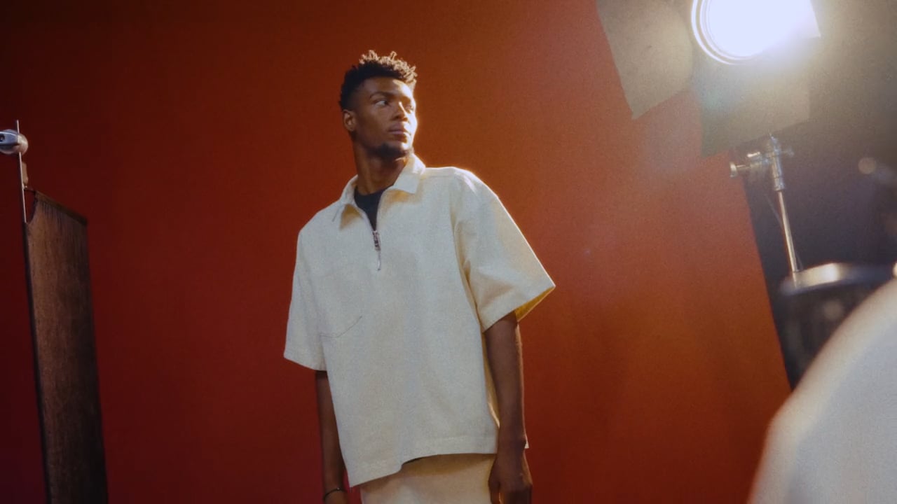 A young adult male poses in a studio setting with soft lighting, wearing a beige short sleeve shirt and matching pants against a red backdrop.