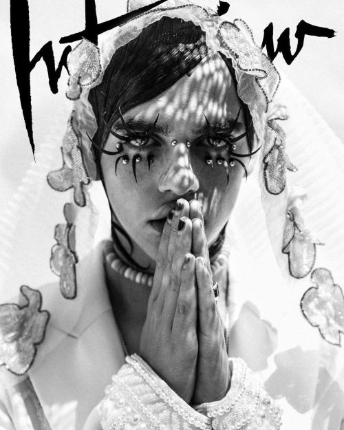 Monochrome portrait of a person with dramatic makeup, wearing a decorative headpiece and pearl necklace, with hands folded in prayer.