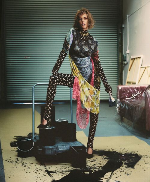 Stylish adult female model posing in patterned top and leggings next to paint cans, with an industrial door in the background.