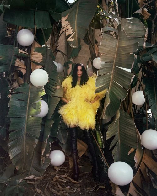 A fashionable woman in a yellow feather coat and black boots poses among lush green banana leaves and glowing white sphere decorations.