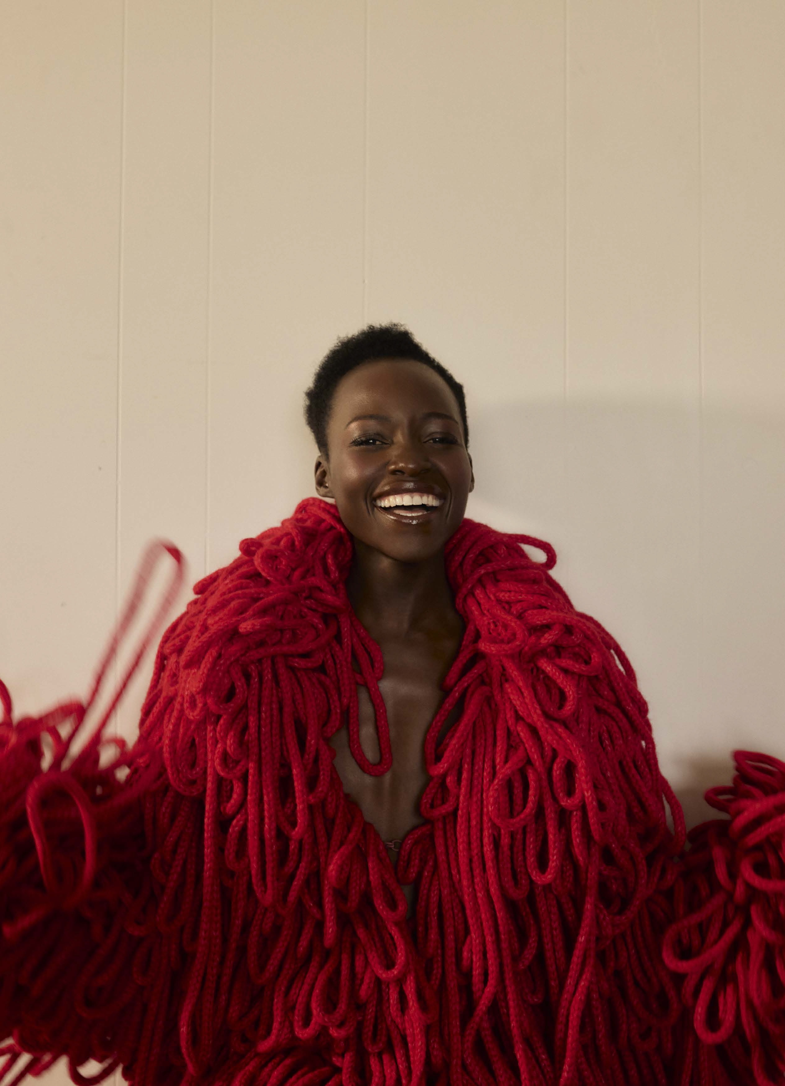 Woman smiles in red yarn coat in front of plain wall. Striking texture and fashion statement. Beauty shot with dark-skinned adult female.