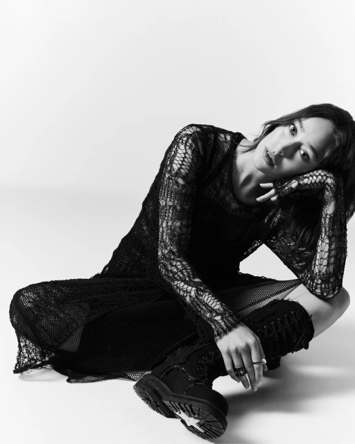 A young adult woman poses in a black lace dress and combat boots, sitting on a white floor with a soft, natural light on her face.