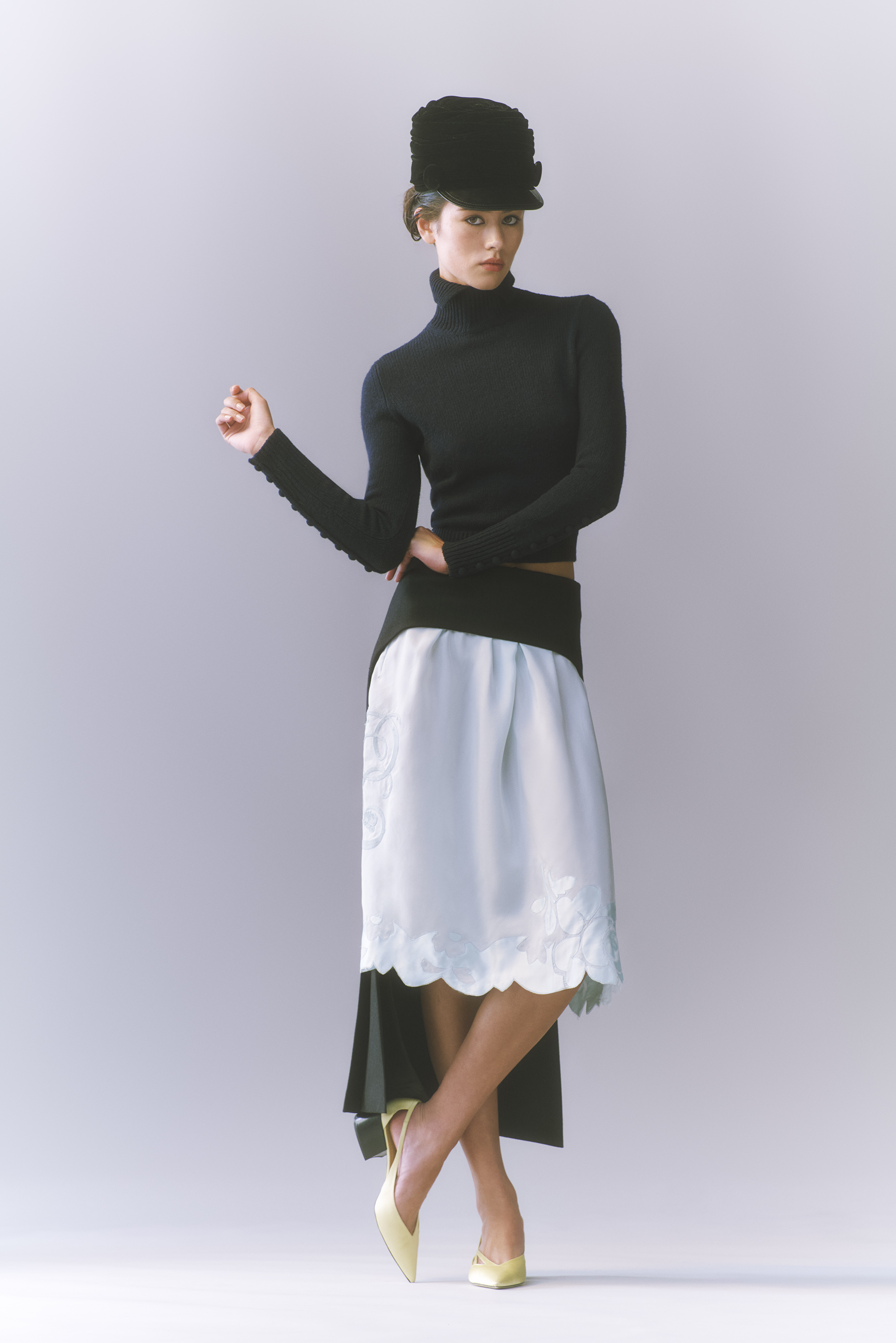 Full-shot of a woman in a black turtleneck, light blue skirt with floral embroidery, black hat, and light yellow heels against a grey background.