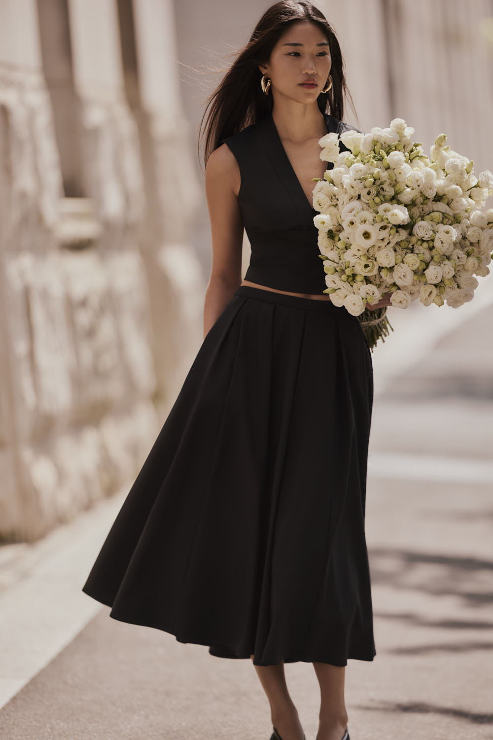 Elegant Asian woman in black dress holds a white floral bouquet outdoors against a neutral building backdrop, complemented by hoop earrings.