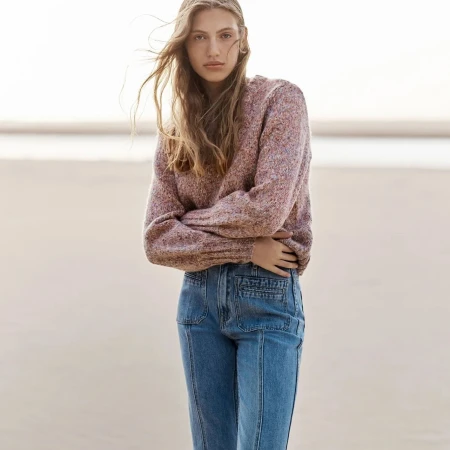 A female model with long blonde hair blowing in the wind, wearing a textured mauve sweater and blue denim jeans against a sandy beach.