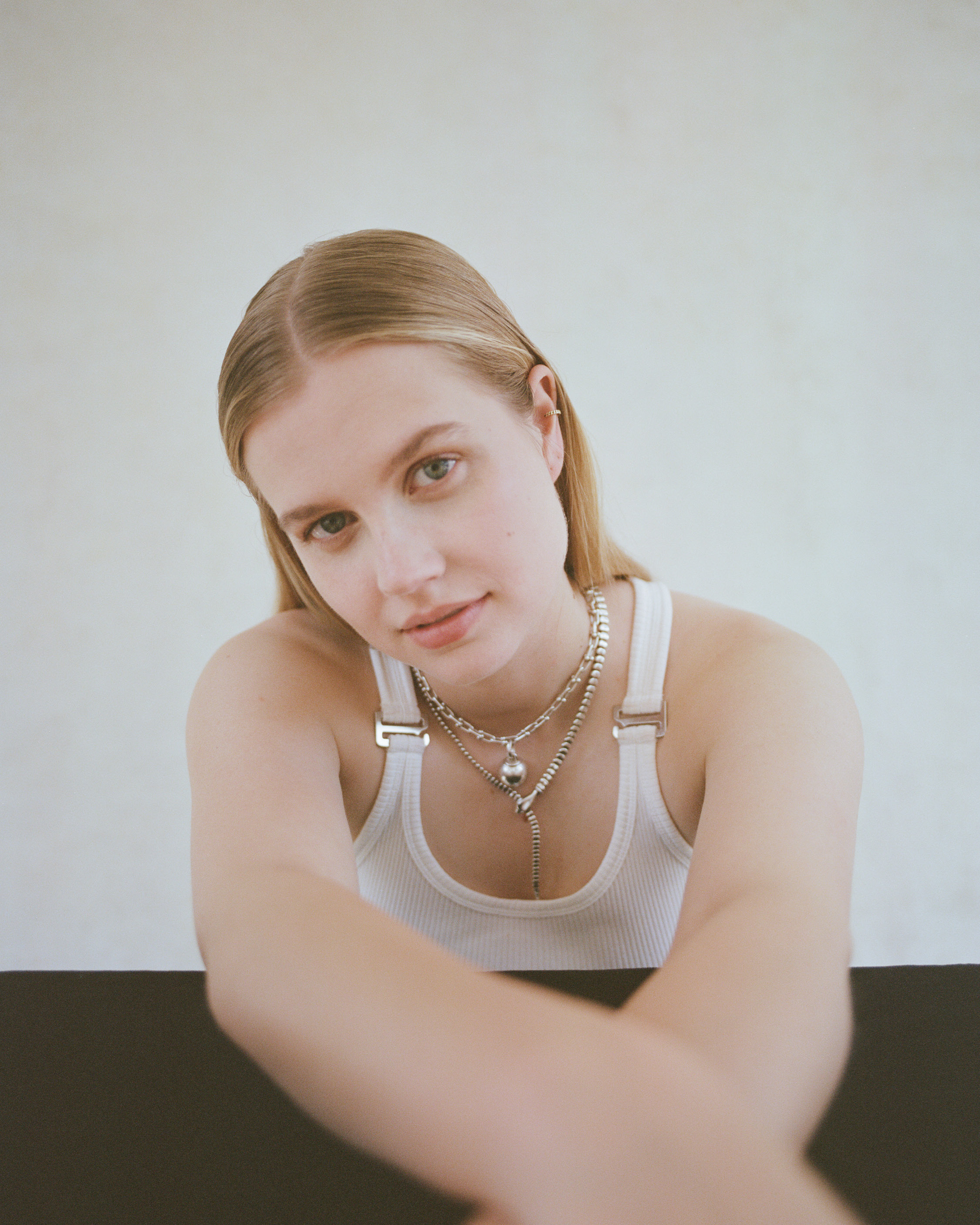 Portrait of a young blonde woman in a white tank top, wearing silver necklaces against a neutral background, arms crossed in the foreground.