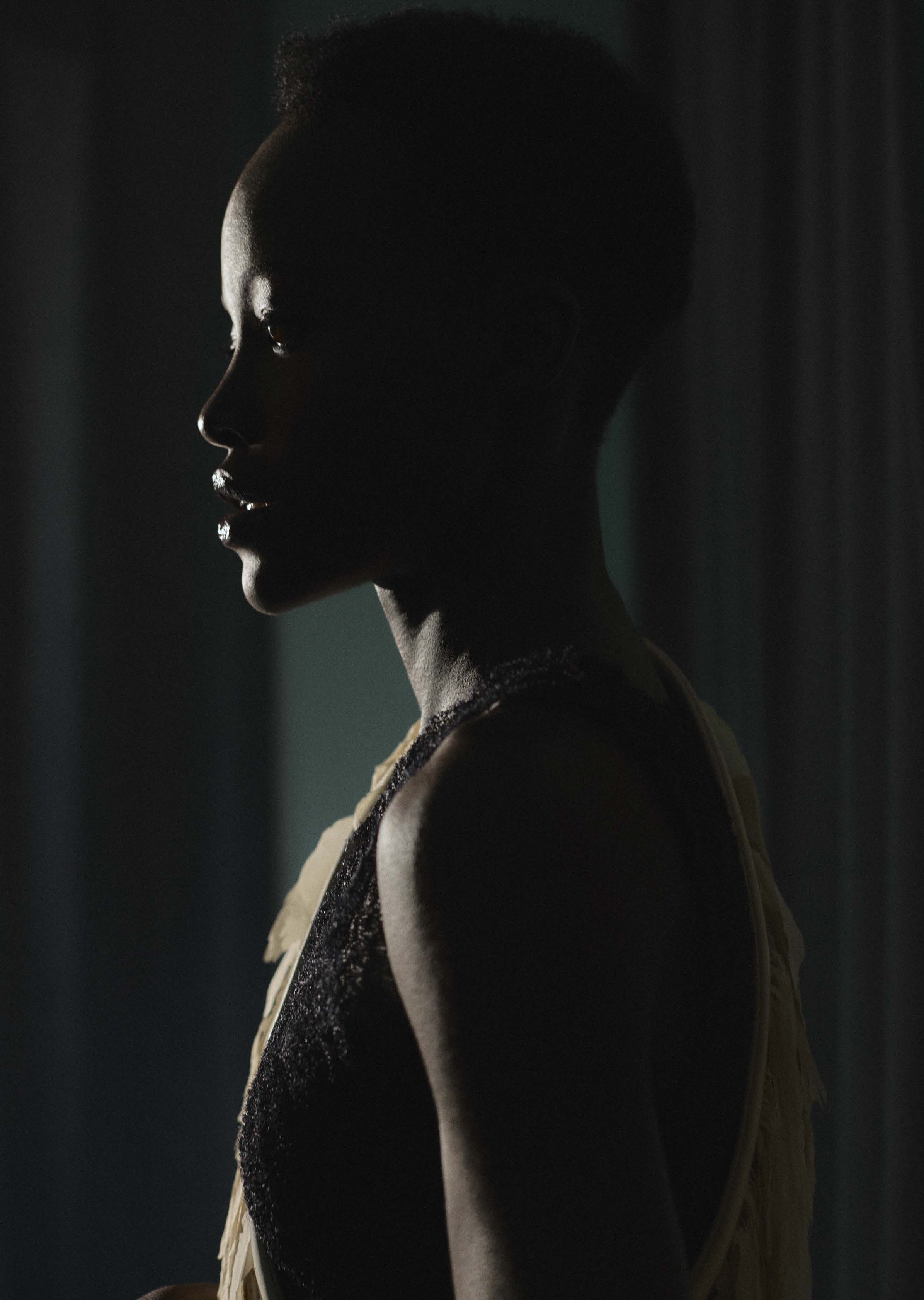 Striking profile of a Black woman with short hair, adorned in a sleeveless dress with textural details, against a softly lit, draped backdrop.