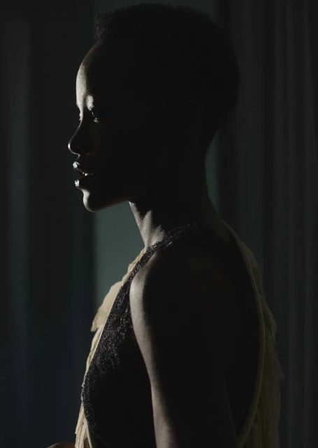 Striking profile of a Black woman with short hair, adorned in a sleeveless dress with textural details, against a softly lit, draped backdrop.