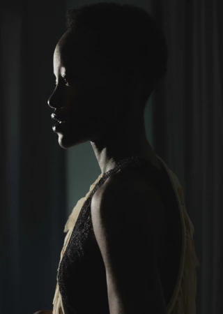 Striking profile of a Black woman with short hair, adorned in a sleeveless dress with textural details, against a softly lit, draped backdrop.
