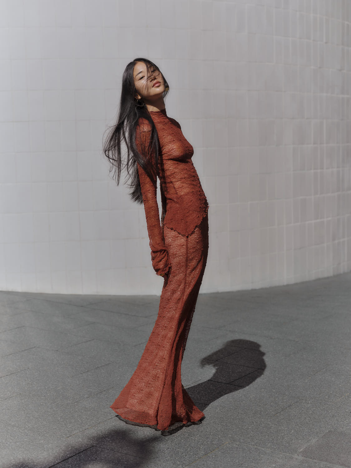 Woman with long dark hair wearing a rust-orange lace top and matching skirt standing outdoors against a curved white wall