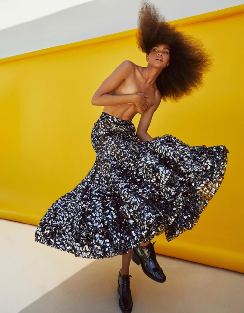 A person with a large afro hairstyle poses in a silver and black skirt and black patent leather shoes in front of a yellow background.