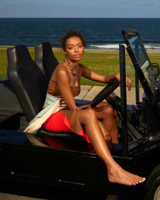 Young woman posing in an open black vehicle on a sunny day, with the ocean in the background, wearing fashionable clothing and gold necklace.