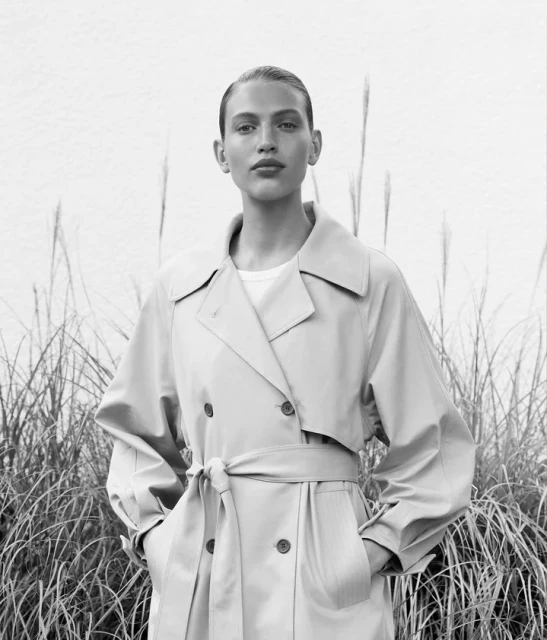 Portrait of a female model in a trench coat, standing outdoors against a light wall and tall grasses, in a fashion style photo shoot.
