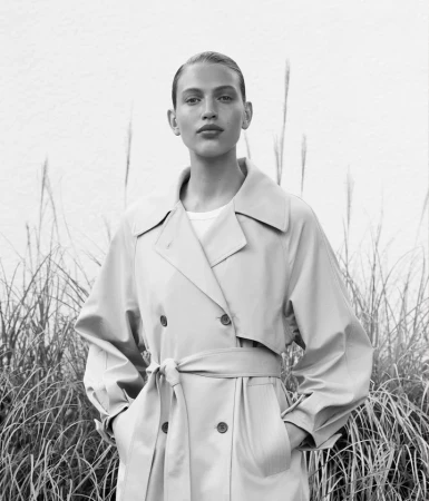 Portrait of a female model in a trench coat, standing outdoors against a light wall and tall grasses, in a fashion style photo shoot.