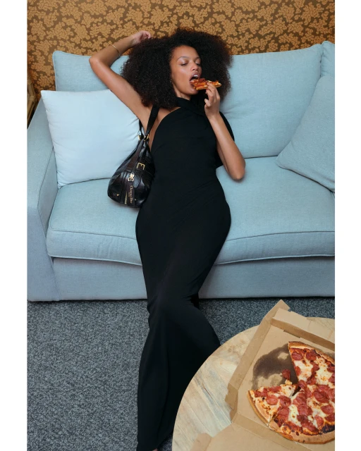 Woman in black dress eats pizza on a couch with a purse, against a wallpapered wall and gray carpet in an indoor setting.