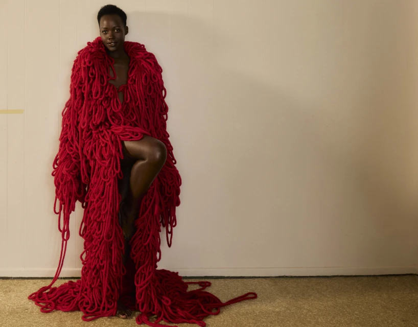 A Black woman poses indoors, draped in a large red yarn garment, with one leg raised and a neutral facial expression.