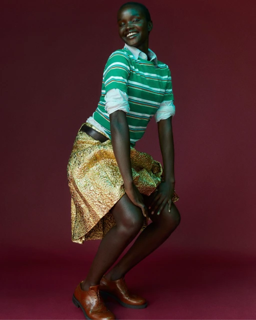 Smiling African woman in a green striped shirt and gold skirt against a red background, wearing brown shoes and teal makeup.