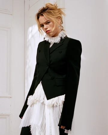 Woman with blonde hair poses in a black blazer with a frilled white collar and a pleated skirt against a white wall in a studio.