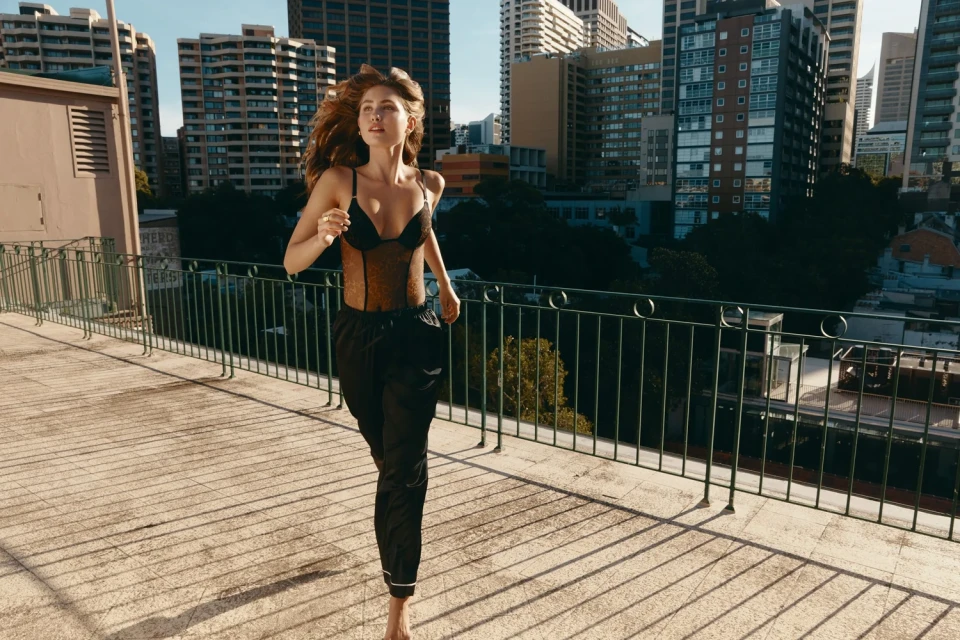 A woman in a black bodysuit and jogging pants runs barefoot on a rooftop overlooking a cityscape with high-rise buildings.