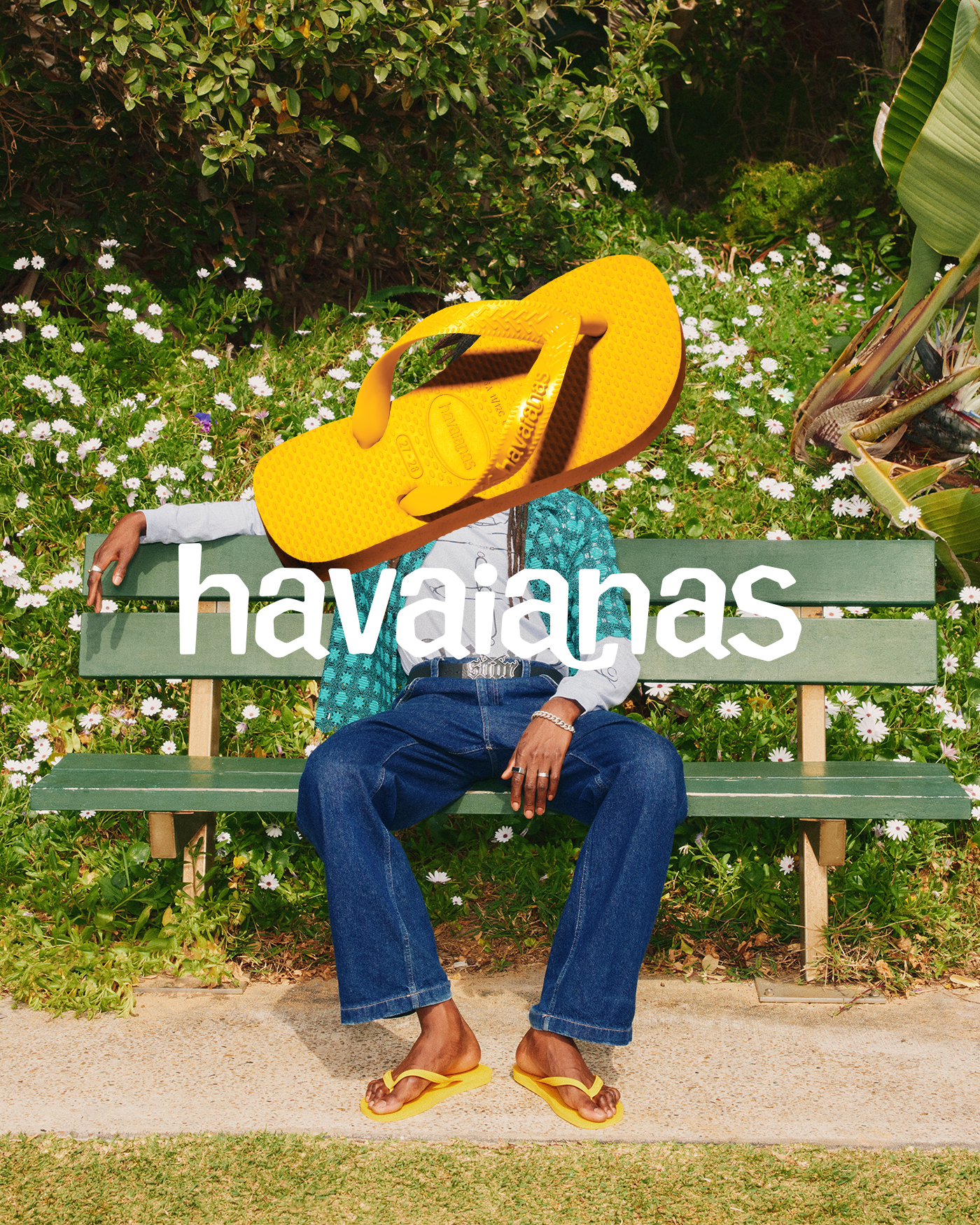 Person sitting on a bench in a park wearing Havaianas yellow flip-flops with a giant yellow Havaianas flip-flop over their head.