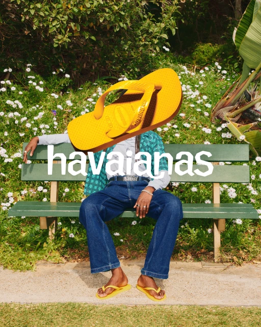 Person sitting on a bench in a park wearing Havaianas yellow flip-flops with a giant yellow Havaianas flip-flop over their head.