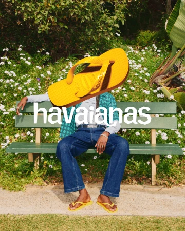 Person sitting on a bench in a park wearing Havaianas yellow flip-flops with a giant yellow Havaianas flip-flop over their head.