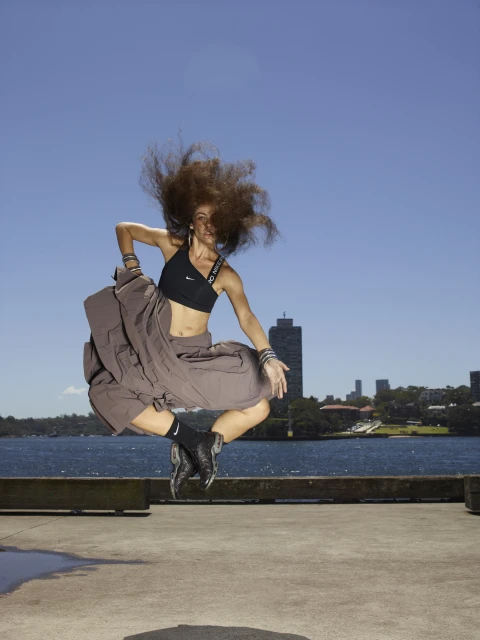 Athletic woman mid-air, leaps gracefully on a concrete platform by the water, backed by a cityscape under a clear blue sky. 