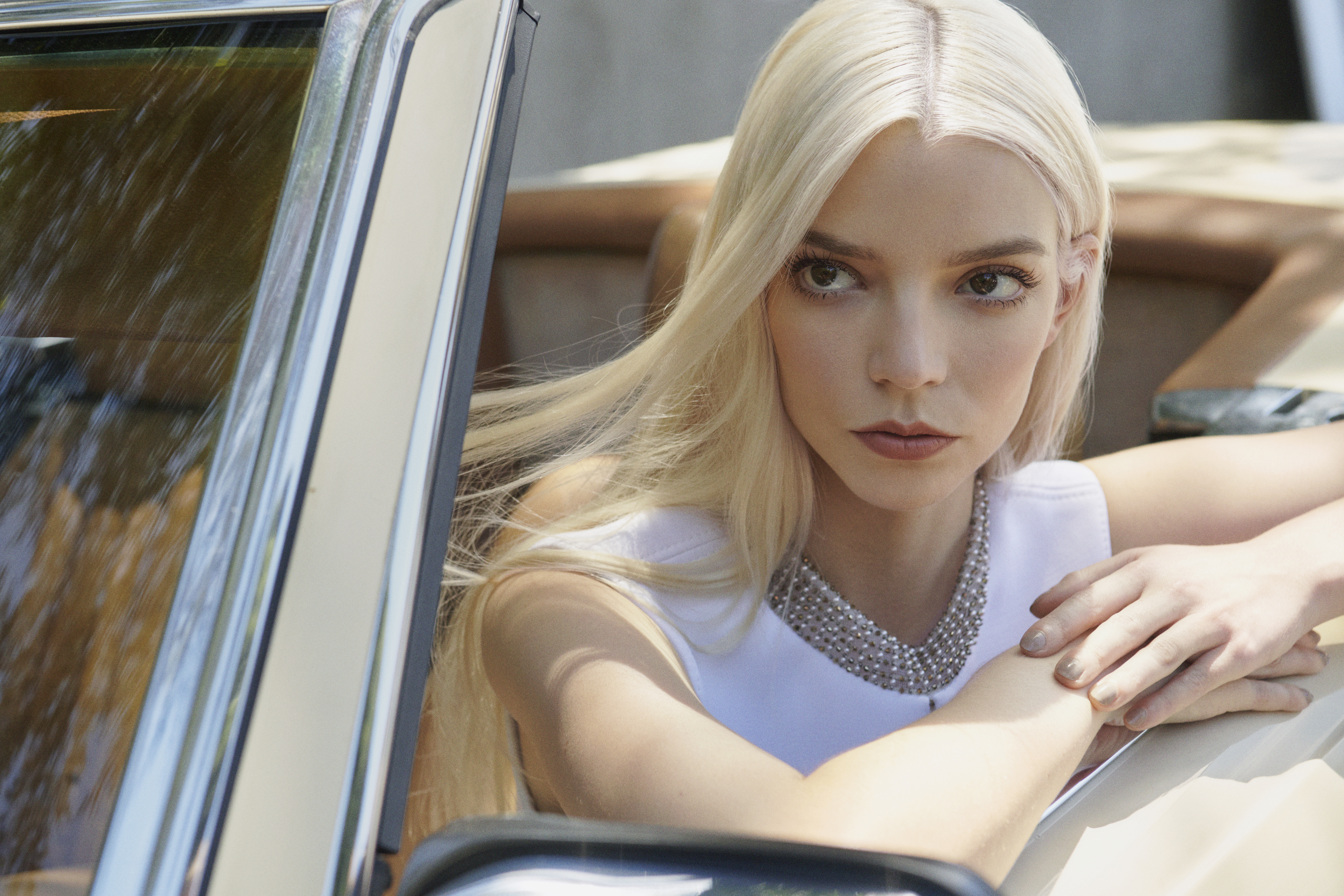 Anya Taylor-Joy in a white dress and silver necklace leans against a vintage car door, her blonde hair falling across her shoulders.